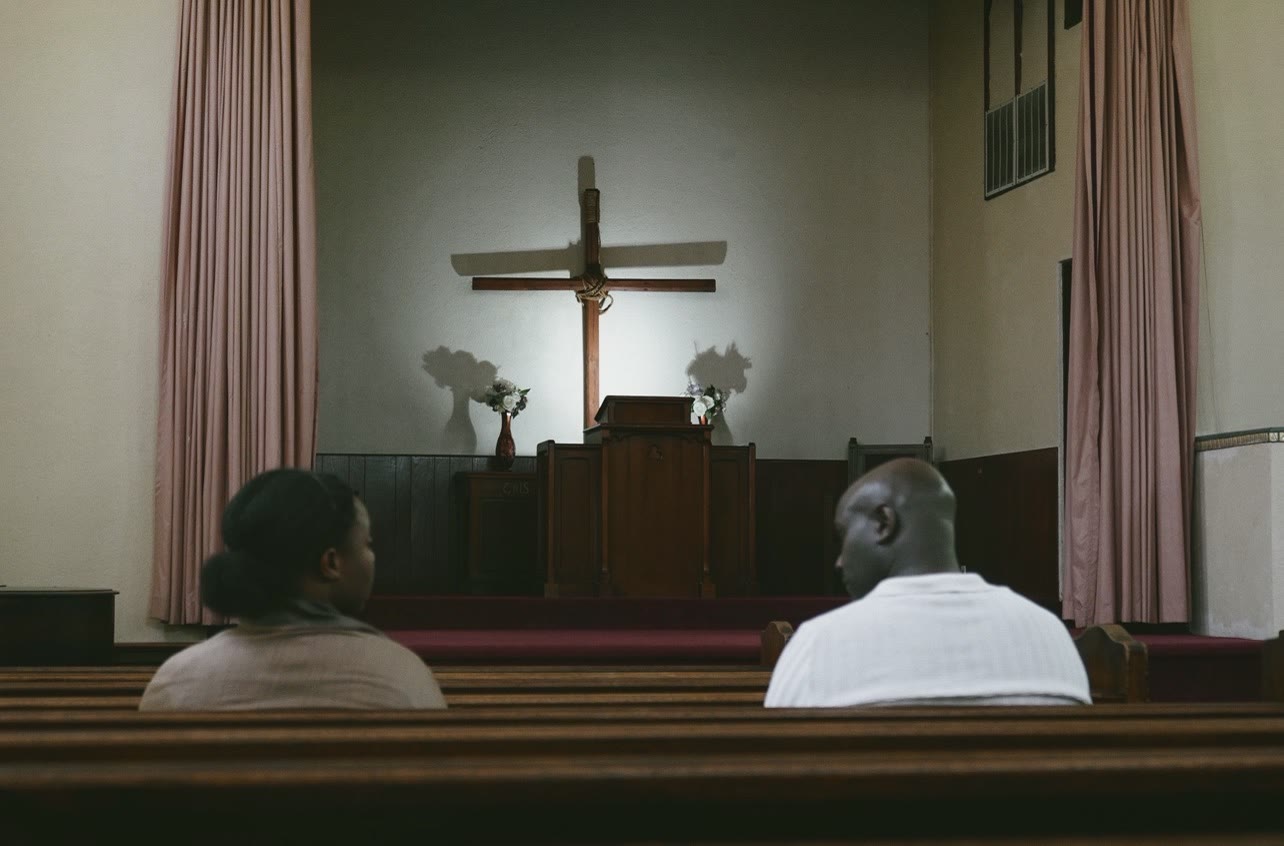 Still from FADING — a couple seated at a table with a cross